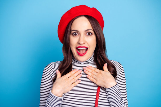 漂亮女孩的照片肖像，惊讶地微笑，双手放在胸前，戴着贝雷帽，穿着条纹高领衫，背景为明亮的蓝色。图片下载