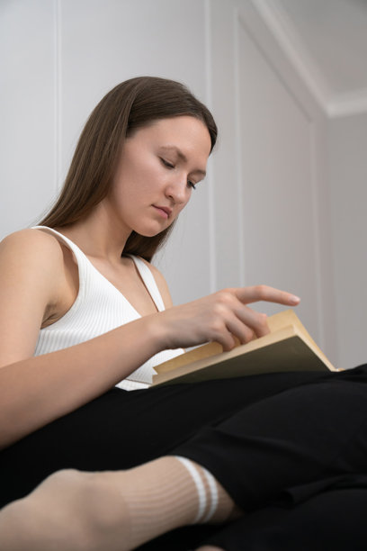 年轻女性穿着袜子和休闲服，舒适地坐在家里的沙发上，享受在自然光下阅读一本平装书。竖版照片图片下载