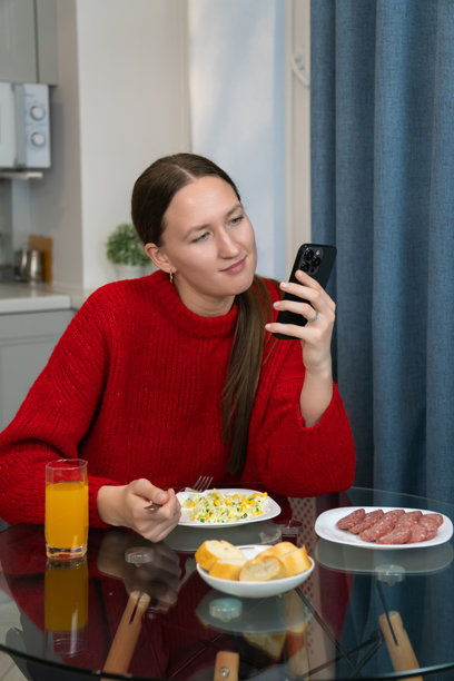 年轻女性穿着红色毛衣，在现代厨房里一边吃早餐一边使用智能手机，享受健康的沙拉、面包、香肠和橙汁。竖版照片图片下载