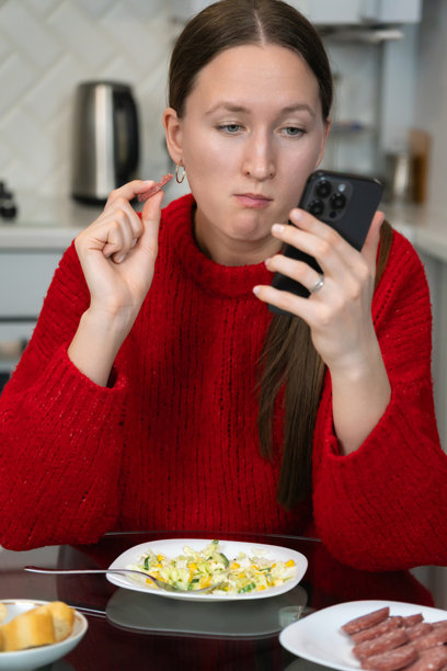 年轻女性穿着红色毛衣，在现代厨房享用早餐，一边吃着一片香肠，一边查看智能手机，享受轻松舒适的早晨日常。竖版照片图片下载