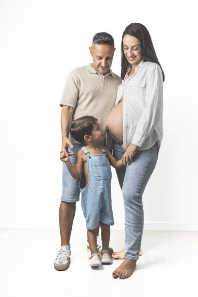 快乐的孕妇与丈夫和可爱的三岁儿子，三人都穿着牛仔裤和浅色衬衫，背景为白色。图片下载