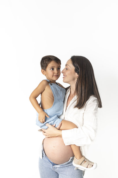 快乐的孕妇怀孕8个月，穿着白色衬衫和牛仔裤，怀抱着可爱的3岁儿子，儿子穿着牛仔工装，背景为白色。图片下载