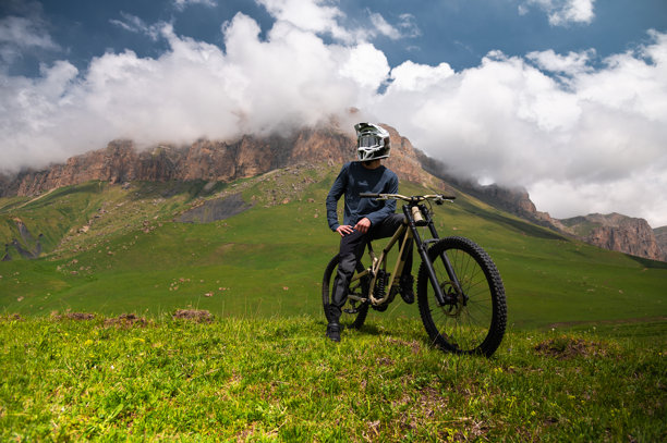 骑自行车的运动员戴着头盔，站在山地车上休息，准备下坡，背景是壮丽的山脉和风景。山地自行车下坡运动的场景。图片下载
