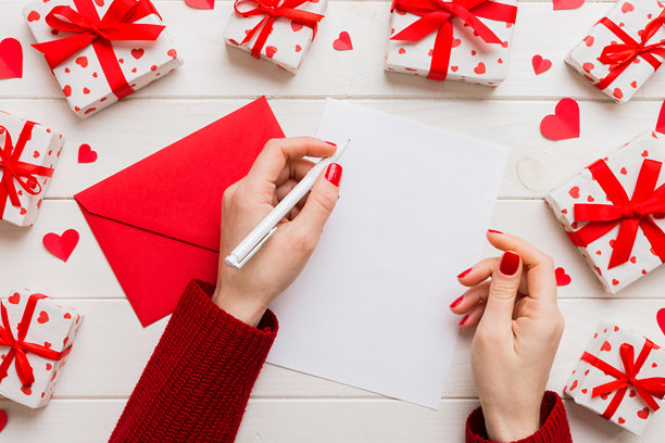 从上方拍摄的情人节装饰照片，女性手持信件和信封，旁边有小礼物盒和心形装饰，背景为单色柔和色调，留有空白空间。图片下载
