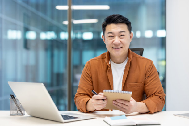 这张照片捕捉到了一位快乐的亚洲男子在桌子旁微笑着拿着一本笔记本。图片下载