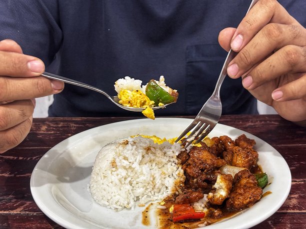 这张图片展示了一个人在吃东西。他的双手各持一把勺子和一把叉子。盘子里有白米饭，一道用深棕色酱汁覆盖的鸡肉或鱼肉块，以及绿色和红色的甜椒片。图片下载