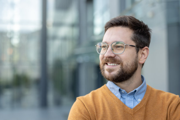 年轻商人在商业中心附近的街道上微笑着侧望，特写照片，留着胡子，戴着眼镜。图片下载