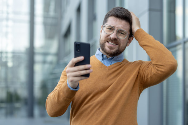 年轻的压力山大的商人在办公室大楼外的特写照片，他手里拿着手机，手托着头，悲伤地看着镜头。图片下载