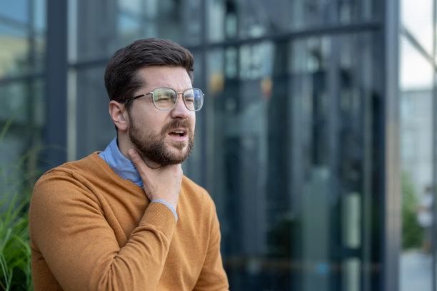 年轻男性办公室工作人员坐在建筑外，手握喉咙，感到剧烈疼痛和炎症的特写照片图片下载