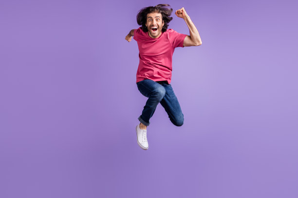 全身照片，乐观的棕发男孩穿着粉色T恤和牛仔裤在紫色背景上跳跃。图片下载