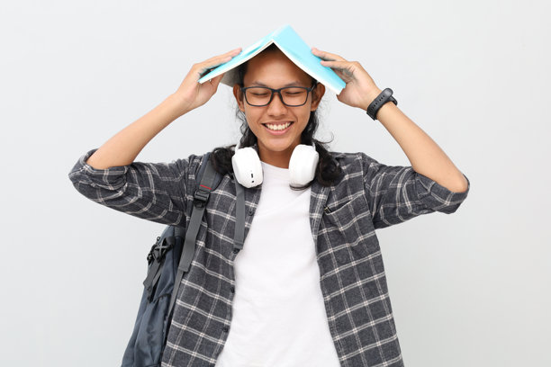 快乐可爱的年轻亚洲男学生，穿着休闲服，背着书包，戴着眼镜，脖子上挂着耳机，微笑着闭上眼睛，手里拿着一本打开的书放在头上。图片下载