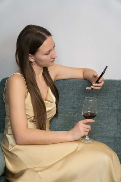 优雅的女性穿着金色缎面裙子，坐在沙发上，使用智能手机，享受一杯红酒，营造出一种精致而放松的氛围。竖版照片图片下载