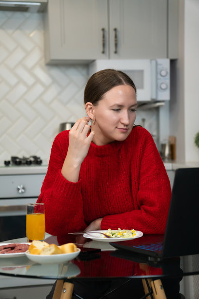 年轻女性穿着红色毛衣，在现代厨房享用早餐，同时在笔记本电脑上浏览互联网，结合了早晨的日常活动与在线活动。竖版照片图片下载