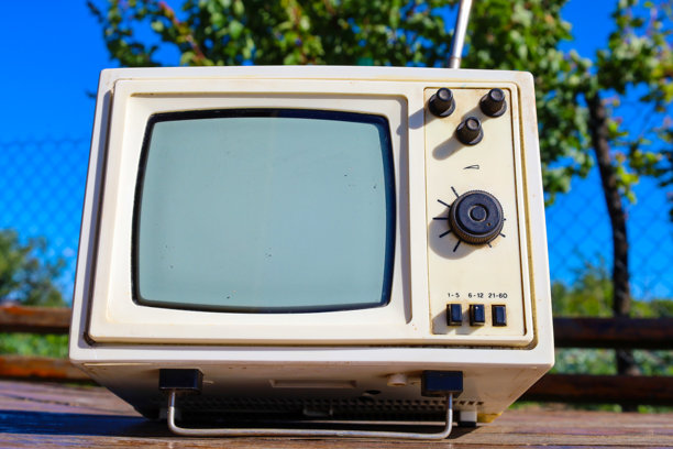 一台黑白屏幕的便携复古电视放在木桌上，背景模糊的绿树和蓝天。这个画面营造出怀旧的感觉，展示了科技的演变。图片下载