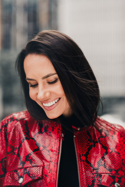 身穿红色印花夹克的时尚年轻女性在阳光明媚的城市户外微笑，竖版照片图片下载