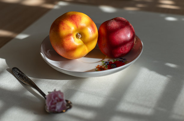 特写照片：两个成熟的桃子（黄色和红色）放在花卉碗中，桌子上有勺子和粉色花朵。图片下载