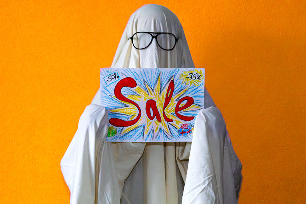 幽默的图像，描绘了一只穿着白色床单服装的幽灵形象，手持红色促销标志。背景为橙色，这张概念照片将恐怖的万圣节意象与零售促销和视觉幽默相结合。图片下载
