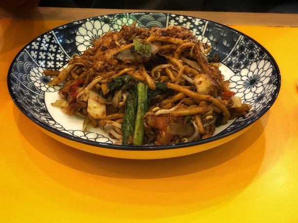 这张图片展示了一盘炒面（米干）放在明亮的黄色桌子上。盘子上有蓝白相间的装饰图案。图片下载