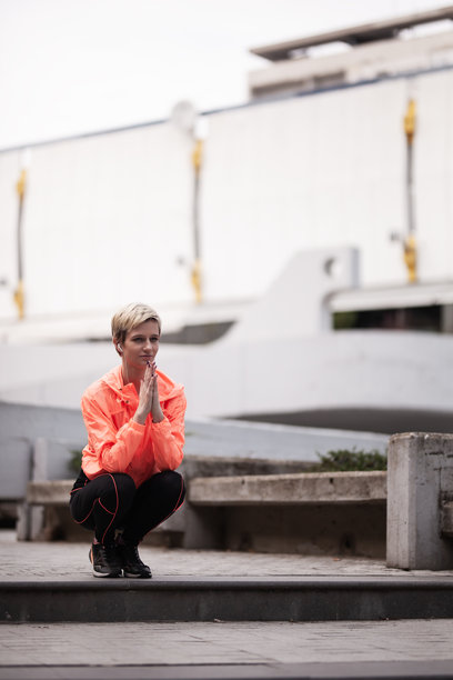 这张图片捕捉到一位年轻女性，穿着明亮的橙色夹克，在城市晨跑时以冥想姿势享受宁静的休息。图片下载