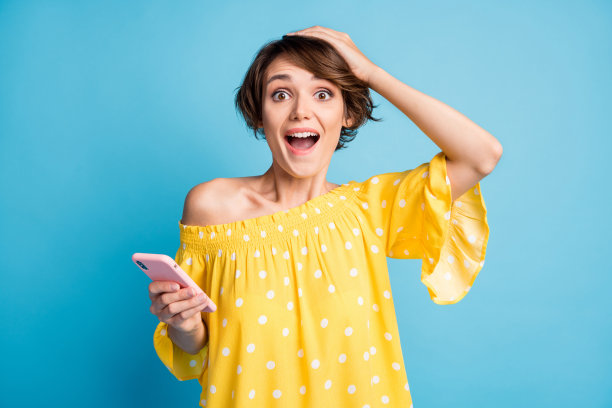 兴奋女孩的照片肖像，单手握着手机，触摸头部，背景为鲜艳的蓝色。图片下载