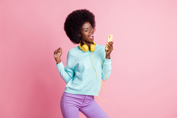 女孩在粉色背景下戴着耳机，手持智能手机跳舞并唱歌的照片肖像图片下载