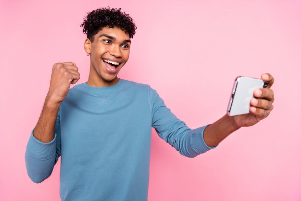 年轻男孩在粉色背景下玩手机游戏，做出胜利手势的照片肖像图片下载
