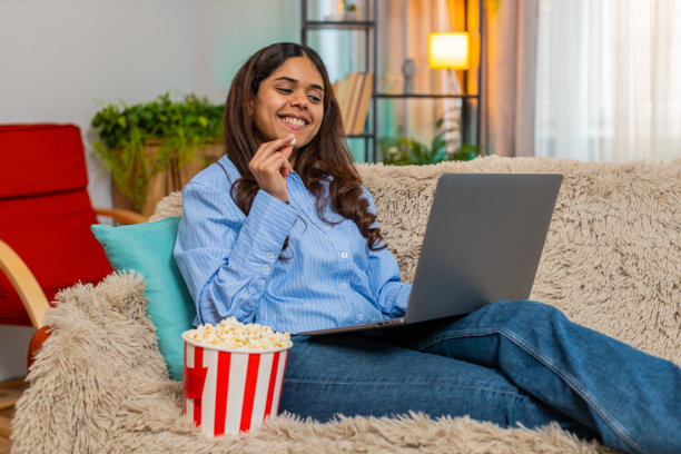 印度女性在家里的沙发上看电影，微笑着看着笔记本电脑屏幕，手里拿着爆米花，享受温馨时刻。图片下载