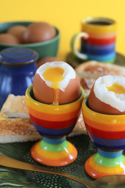 早餐场景的图片，绿色向日葵设计的盘子，两个彩虹条纹的蛋杯里盛着流心的软煮蛋，白色涂黄油的吐司士兵，金属茶匙，蓝色盐罐，一碗鸡蛋，彩虹条纹的浓缩咖啡杯，黄色背景。图片下载