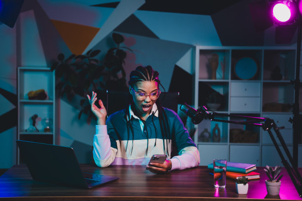 迷人的女性博主在时尚的室内办公室环境中，坐在桌子前录制播客，周围有明亮的霓虹灯光。图片下载