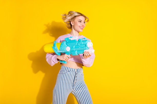 年轻快乐女孩的侧面照片，面带微笑，手持玩具枪，背景为黄色。图片下载