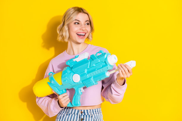 快乐、开朗、漂亮的年轻女性在黄色背景下玩水枪，面带微笑，目光投向空旷的空间。图片下载