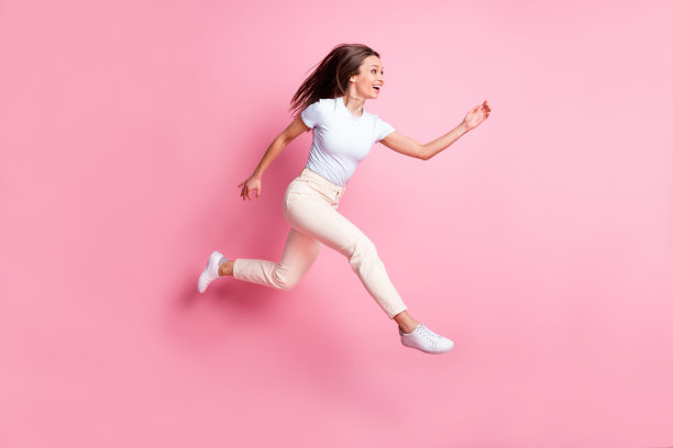 乐观的棕发女士穿着T恤和裤子在粉色背景上跳跃的全身侧面照片图片下载