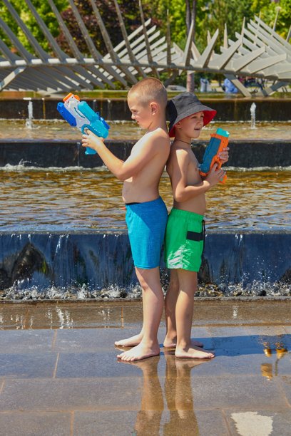 水上对决。两个穿着泳衣的男孩在阳光明媚的日子里，在喷泉附近玩水枪。图片下载