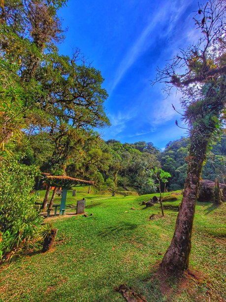 巴西巴拉那州莫雷特斯的格拉西奥萨大道上的卡斯卡塔河休闲区。照片展示了野餐桌和一个庇护所，周围环绕着保存完好的大西洋森林，绿草如茵，大石头和热带树木在蓝天之下。图片下载