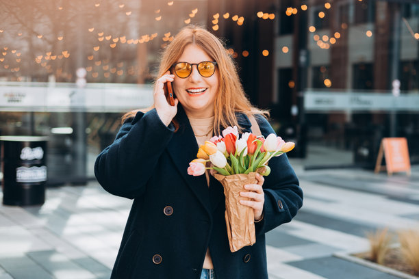 微笑的女性手持郁金香在户外打电话。快乐的女孩在户外摆姿势。春季城市背景下美丽女性的肖像。高质量照片。图片下载