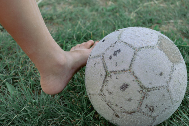 一只脚靠近一只旧足球，站在绿色草地上的特写镜头捕捉了儿童游戏和夏季休闲的精神。这张照片非常适合展示运动、童年、积极休闲和户外游戏的主题。图片下载