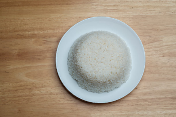 平铺视角拍摄的蒸熟泰国茉莉香米，放在干净的白色盘子里，孤立在木质餐桌的一角，照片留有空白空间供文字使用。图片下载