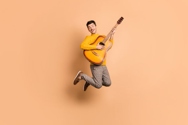 全身肖像照片，快乐的男孩手持吉他在米色背景上跳跃图片下载