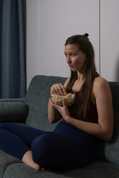年轻女性坐在家里的沙发上，认真专注地享受着从玻璃碗里取出的爆米花，沉浸在电影或电视节目中。竖版照片图片下载