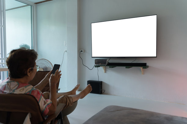 老年女性在现代客厅中放松，观看空白的电视屏幕，同时使用智能手机。图片下载