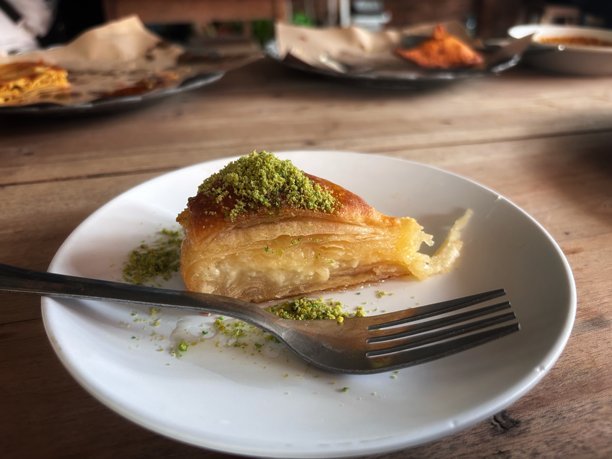 这张图片展示了一块放在白色盘子上的巴克拉瓦。糕点有金黄色的酥皮层，上面撒有切碎的绿色开心果。旁边放着一把叉子。背景中的木桌营造出温暖的乡村氛围。图片下载