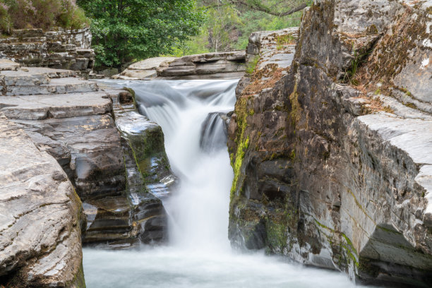 长时间曝光的瀑布照片，瀑布位于覆盖着地衣和苔藓的岩石之间，背景是树木和石南花。奎奇峡谷。图片下载