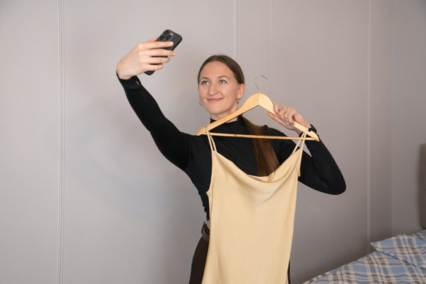 时尚博主在卧室里拿着米色吊带裙和木衣架，用智能手机拍照，为她的时尚博客或社交媒体创作内容。图片下载
