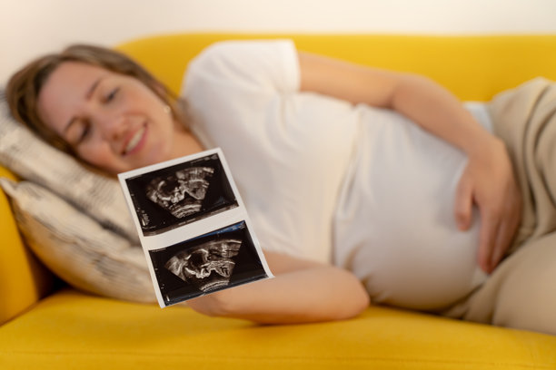 一位快乐的准妈妈坐在沙发上，满怀期待地握着她的超声波照片。图片下载