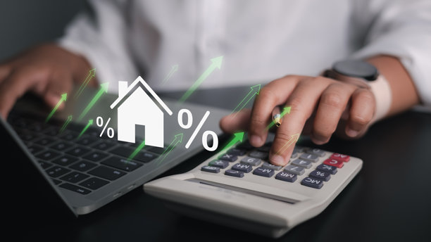 一个人使用笔记本电脑和计算器，旁边漂浮着全息房屋图标和百分比符号，代表着房地产价值上升、抵押贷款利率或物业投资与财务增长。图片下载
