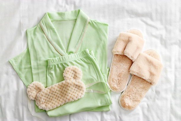 俯视折叠的浅绿色女性睡衣、毛绒睡眠眼罩、柔软的家居拖鞋，摆放在白色床单上。极简风格的舒适睡衣平铺。时尚睡衣的美学生活照片。图片下载