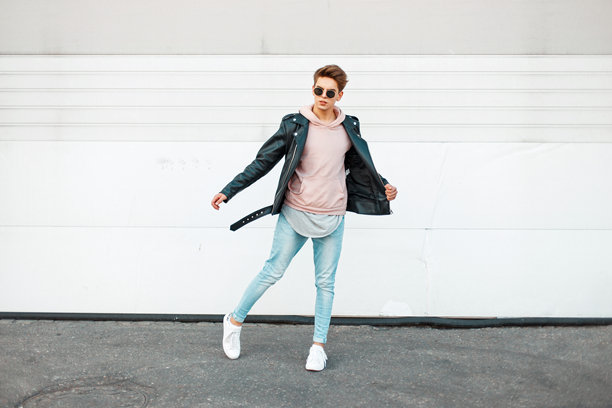 身穿黑色皮夹克、粉色卫衣、蓝色品牌牛仔裤和白色鞋子的时尚帅气年轻男子在运动中图片下载