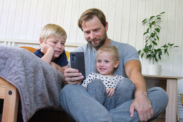 年轻爸爸和孩子们一起使用智能手机。男人在手机上观看内容。快乐的父亲、女儿和儿子在看搞笑视频时开心地微笑，假期的照片，玩手机游戏。图片下载