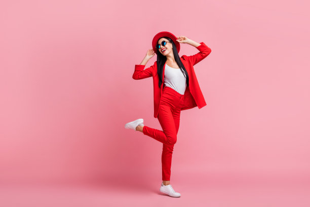 年轻漂亮女孩的全身照片，开心地微笑，双手触摸帽子，目光投向空旷的地方，背景为粉色。图片下载