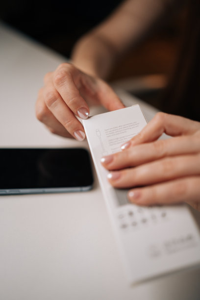 垂直裁剪的照片，女人正在打开装有新手机线缆适配器的白色零售盒，手握盒子，旁边是一张干净的白色桌子，上面放着智能手机。图片下载